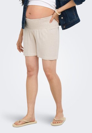 Femme enceinte portant un short beige à taille élastique, un débardeur blanc, une veste en jean et des tongs beiges, debout avec la main sur la hanche.