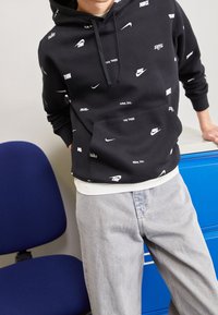 Sudadera con capucha negra con logotipos blancos de Nike dispersos, ajuste holgado, bolsillo tipo canguro. Combinada con jeans gris claro, tela texturizada.