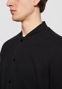 Svart snap-knapp-jakke med ståkrage, laget av teksturert stoff. Har et minimalistisk design uten synlige mønstre eller akenter.