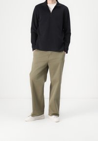 Haut à manches longues en bleu marine foncé avec fermeture éclair, associé à un pantalon large couleur olive clair et des baskets blanches. Design simple et fonctionnel avec des textures douces.