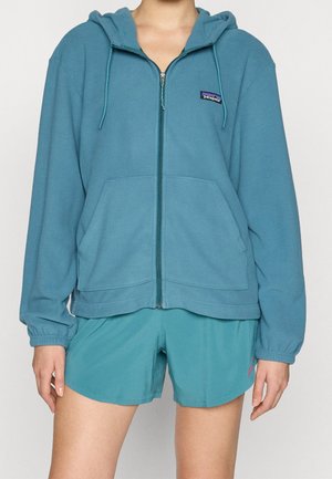Henkilö, jolla on päällään petroolinvärinen vetoketjullinen fleece-huppari, jossa on Patagonia-logo, sekä vastaavat petroolinväriset urheilushortsit yksiväristä taustaa vasten.