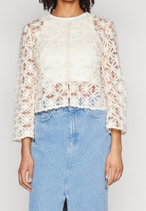 White, sheer floral embroidered cropped jacket with long sleeves and a round neckline, paired with a light blue denim skirt.