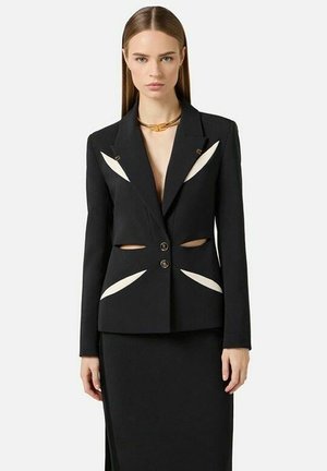 Donna con giacca nera con dettagli bianchi ritagliati e gonna nera abbinata, in piedi davanti a uno sfondo chiaro uniforme.