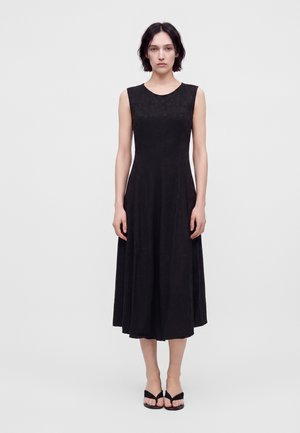 Femme aux cheveux foncés portant une robe midi noire sans manches et des sandales noires slip-on, debout devant un fond gris clair uni.