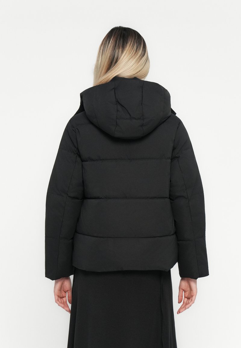Schwarze Pufferjacke mit Kapuze, in boxy Form, mit gesteppten Paneelen und weiten Ärmeln, getragen über einem langen schwarzen Kleidungsstück.