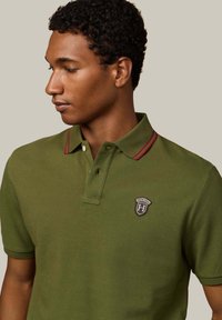 Zelený polo tričko s textúrovaným povrchom, s dvojitým zapínaním na gombíky, golierom s pruhmi v červenej a námorníckej modrej farbe a s logom v tvare štítu na hrudi.