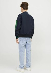 Giubbotto bombardiere navy con dettagli verdi sulle maniche, polsini e vita elasticizzati, indossato sopra jeans azzurri e sneaker bianche.