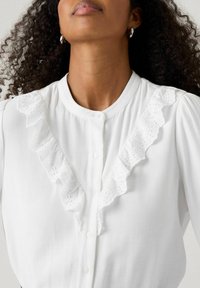 Camicia bianca con bottoni sul davanti, con un pizzo arricciato al centro e tessuto morbido e testurizzato. Collo rotondo e maniche a sbuffo.