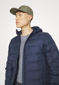 Giubbotto imbottito blu navy con cappuccio, dotato di uno strato base grigio testurizzato sotto e un cappellino verde oliva. Dettaglio del logo discreto sul giubbotto.