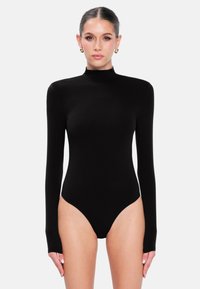 Femme aux cheveux plaqués en arrière, portant des boucles d'oreilles en or en forme de créole et un body noir ajusté à manches longues avec un col montant, se tenant devant un fond blanc.