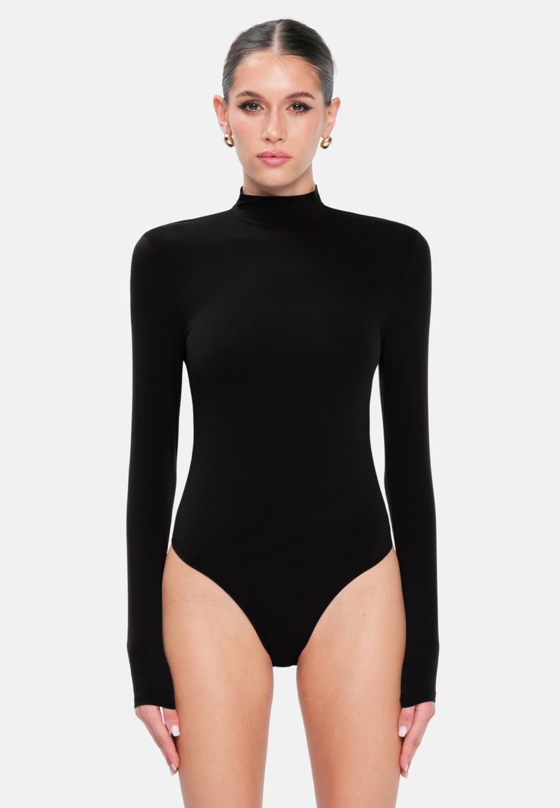 Femme aux cheveux plaqués en arrière, portant des boucles d'oreilles en or en forme de créole et un body noir ajusté à manches longues avec un col montant, se tenant devant un fond blanc.