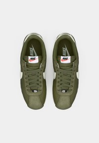 Olīvu zaļas Nike kedas ar baltām akcentiem, mīkstināta mēle un teksturētas šņores. Zole ir viegla; logo uz papēža un mēles.