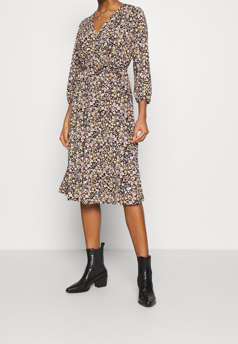 Czarna sukienka w kwiaty z rękawami o długości 3/4, dekoltem w serek i wiązanym pasem. Spódnica ma falbaniasty dół. Dodatkiem są czarne botki za kostkę.