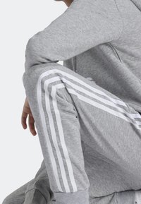 Chándal atlético gris con rayas blancas que bajan por los lados de los pantalones. Textura suave, corte casual, cuello redondo en la parte superior.