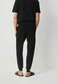 Schwarze Baumwoll-Jogginghosen mit schmal zulaufenden Beinen, elastischen Bündchen, einer Gesäßtasche, kombiniert mit schwarzen Slippern und einem schlichten schwarzen T-Shirt.