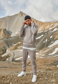 Sudadera beige con bolsillo frontal y logo, combinada con pantalones jogger a juego. Ambas prendas están hechas de una tela suave y se llevan con zapatillas blancas.