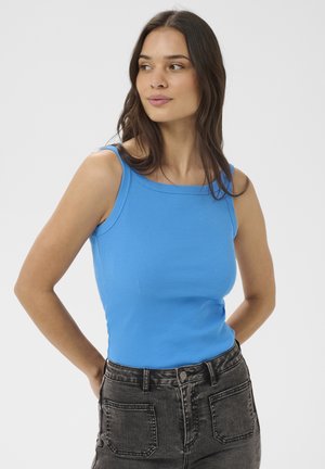 Woman with long brown hair wearing a bright blue sleeveless top and black high-waisted jeans, looking to the side with hands behind her back.
