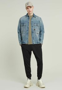 Jeansjacka i ljusblått med två bröstfickor, över en olivgrön t-shirt, tillsammans med svarta joggers och beige sneakers.