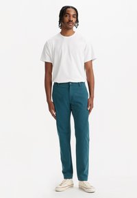 Pantaloni din twill de bumbac teal, cu croială dreaptă, asortați cu un tricou alb simplu cu guler rotund și sneakers din pânză bej.