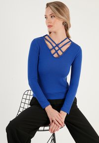 Plavi dugoročan top s rebrastim uzorkom koji ima V-izrez s preklapajućim trakama. Tekstura je glatka, s uskim krojem i minimalističkim detaljima.