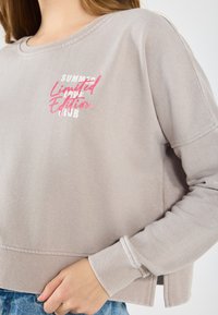 Lys grå genser med rund hals, ribbestrikkede mansjetter og nederkant. Har teksten "SUMMER Limited Edition CLUB" trykt i rosa og hvitt.