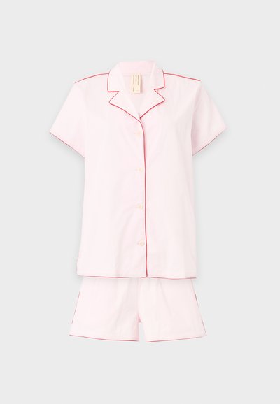 Haut de pyjama à manches courtes rose clair avec boutons et short assorti avec un liseré rouge le long des bords, présenté sur un fond uni.
