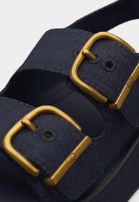 Κοντινό πλάνο από λουριά σανδαλιού σε ναυτικό μπλε χρώμα με υφασμάτινη υφή και δύο χρυσούς ρυθμιζόμενους μεταλλικούς τοκάδες.