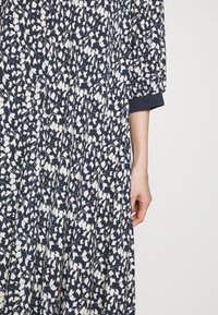 Robe florale présentant un motif bleu marine et crème, avec de longues manches bouffantes dotées de détails de poignet, et un design fluide et plissé.