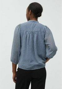 Blouse bleue en tissu léger avec un motif à pois blancs, dos froncé et manches trois-quarts bouffantes. Portée avec un pantalon noir.