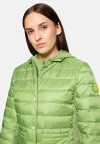 Zielona pikowana kurtka z kapturem, o gładkiej fakturze, z zapięciami na zatrzaski i naszywką z uśmiechniętą buźką na lewej rękawie.