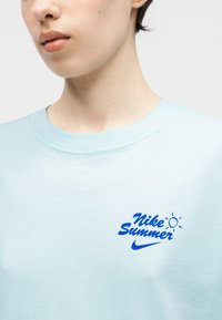 Młoda osoba ubrana w jasnoniebieską koszulkę Nike z niebieskim napisem "Nike Summer" i grafiką słońca na piersi.