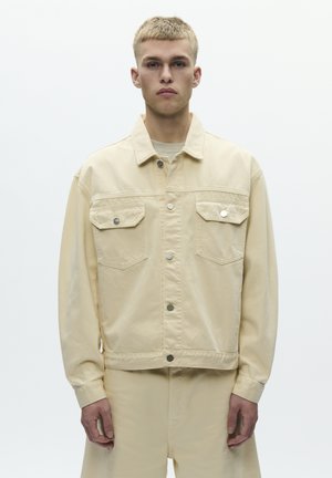 Ung mand med kort blondt hår iført en beige jakke med knapper og matchende bukser foran en ensfarvet hvid baggrund.