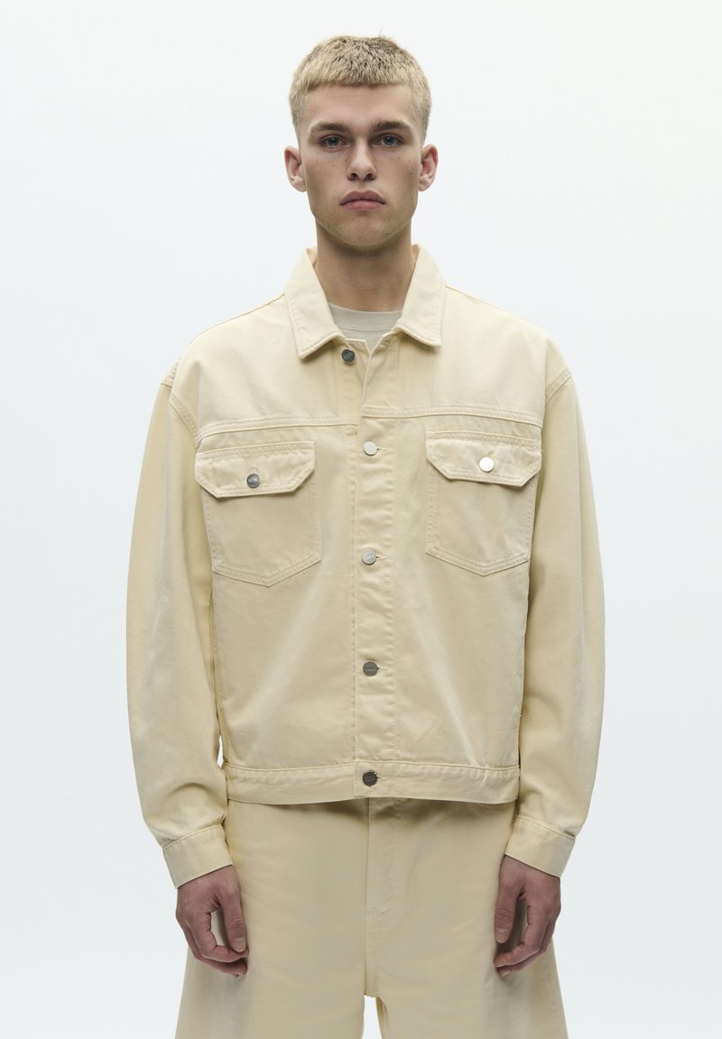 Young man with short blond hair wearing a beige buttoned jacket and matching pants against a plain white background.