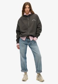 Dunkelgrauer Kapuzenpulli mit Reißverschluss, seitlichen Taschen und Kordelzugkapuze, darüber ein helles rosa Shirt, kombiniert mit hellblauen, hochgekrempelten Jeans und beigen Sneakers.