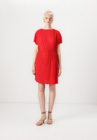 Vestido plissado vermelho com decote arredondado e cintura elástica. Apresenta mangas curtas e cai logo acima dos joelhos. Usado com sandálias beijas de tiras.