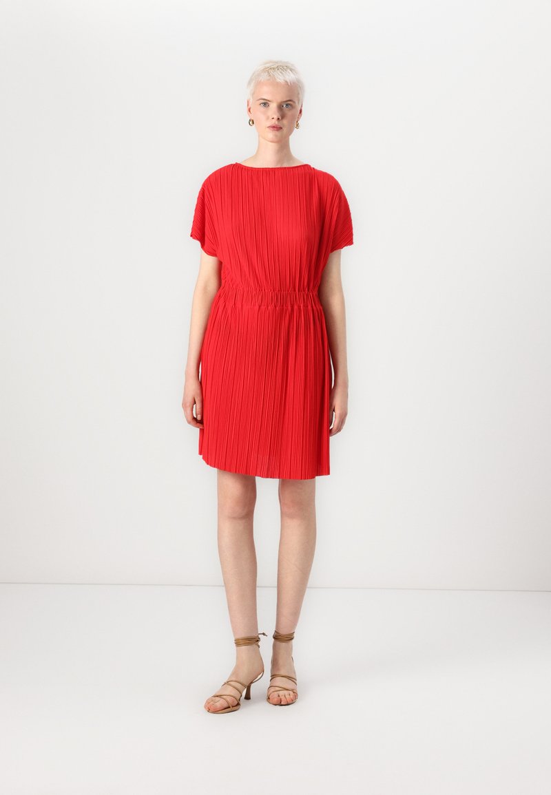 Vestido plissado vermelho com decote arredondado e cintura elástica. Apresenta mangas curtas e cai logo acima dos joelhos. Usado com sandálias beijas de tiras.