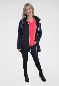 Marinejacke mit weißen Akzenten, Knopfdetails und Kapuze, kombiniert mit einem korallenfarbenen Oberteil und dunklen Jeans, getragen mit schwarzen Stiefeletten.