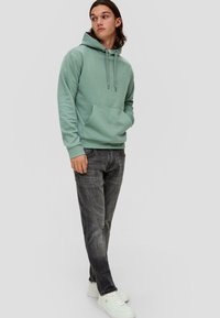Groene hoodie met een kangoeroe-pocket en verstelbare trekkoorden, gecombineerd met donkergrijze jeans en witte sneakers. Casual pasvorm en een zachte textuur.