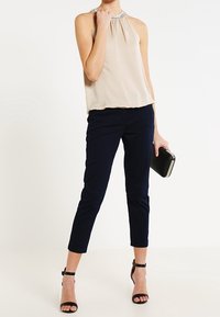 Beige ärmlös topp med en rynkad halsringning och dekoration, kombinerad med figurnära marindrivna byxor som slutar vid anklarna och svarta remmar med klackar.