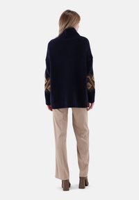 Dunkelblauer Strickpullover mit hohem Kragen, der braune geometrische Muster an den Ärmeln aufweist. Kombiniert mit beigen weit geschnittenen Hosen und braunen Absatzschuhen.