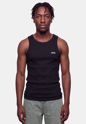 Mann mit geflochtenem Haar, der ein schwarzes ärmelloses Shirt mit der Aufschrift "CRYSTAL" trägt, steht vor einem schlichten hellen Hintergrund.