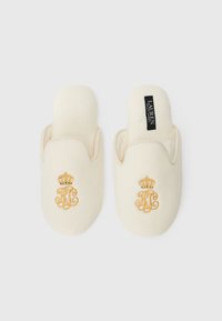 Papuci de culoare crem, realizați dintr-un material moale, cu un emblema brodată în aur, reprezentând o coroana. Design de tip slip-on, cu vârfuri rotunjite și interior pufos.