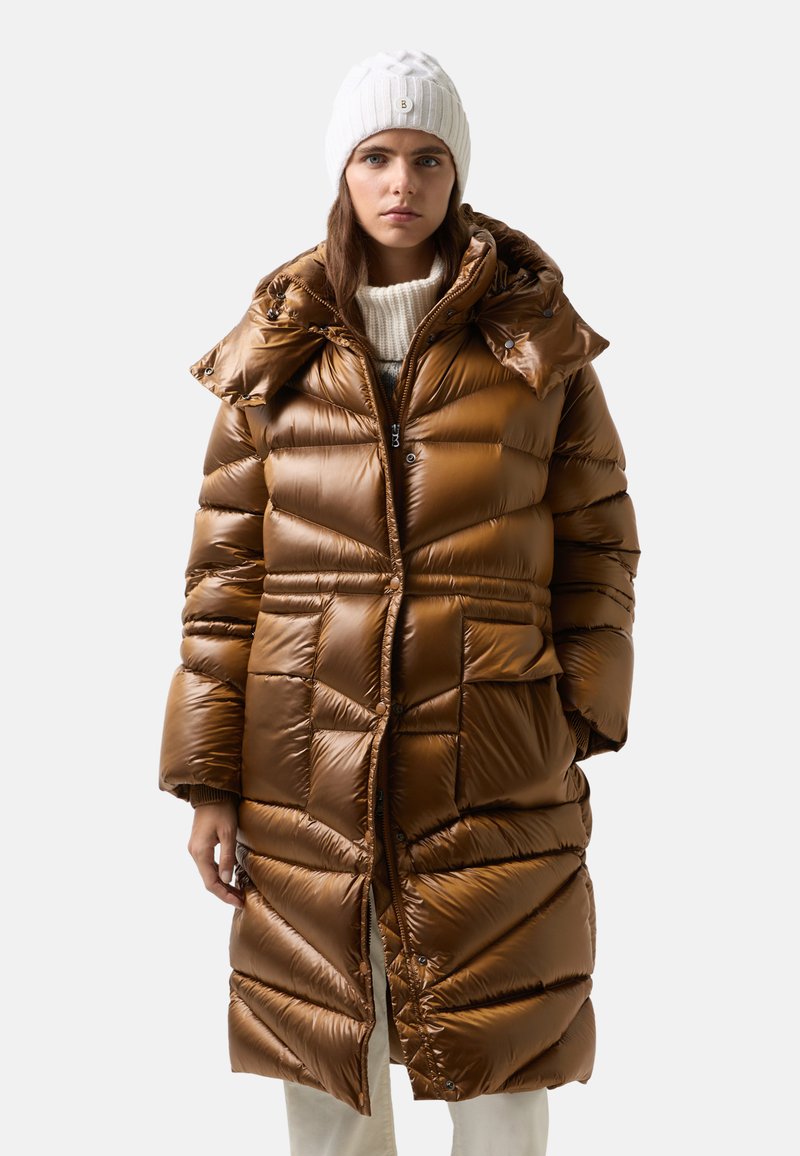 Kobieta w błyszczącym, brązowym, ocieplanym płaszczu, białej wełnianej czapce i kremowym swetrze, stoi z rękami w kieszeniach na jednolitym tle.