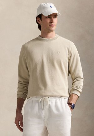 Sudadera beige de manga larga con cuello redondo y puños acanalados. Gorra blanca con logo azul. Pantalones blancos con cordón. Reloj en la muñeca izquierda.