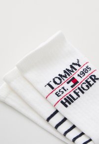 Bela bombažna nogavica z rebrasto teksturo, ki ima logotip "TOMMY HILFIGER" v črni in rdeči barvi. Ena para ima mornarsko modre črte na dnu.