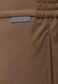 Brūns audums ar smalku rievotā tekstūru, piedāvājot elastīgu jostu un pelēku etiķeti ar tekstu "BDRN" izcili redzamā vietā.