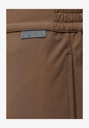 Brūns audums ar smalku rievotā tekstūru, piedāvājot elastīgu jostu un pelēku etiķeti ar tekstu "BDRN" izcili redzamā vietā.