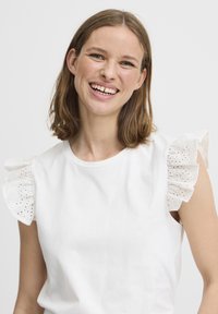 Haut en coton blanc avec des manches courtes à volants en tissu brodé, présentant de petits motifs découpés et un col rond.