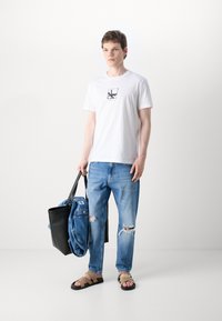 Camiseta blanca de manga corta con un diseño gráfico negro, combinada con jeans rotos de color azul claro y sandalias de tiras marrones, llevando una bolsa tote negra.