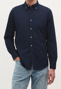 Marineblaue, langärmelige Hemdbluse mit Knöpfen, einer Brusttasche und einem geknöpften Kragen; kombiniert mit hellblauen Jeans. Glatte Stoffstruktur.